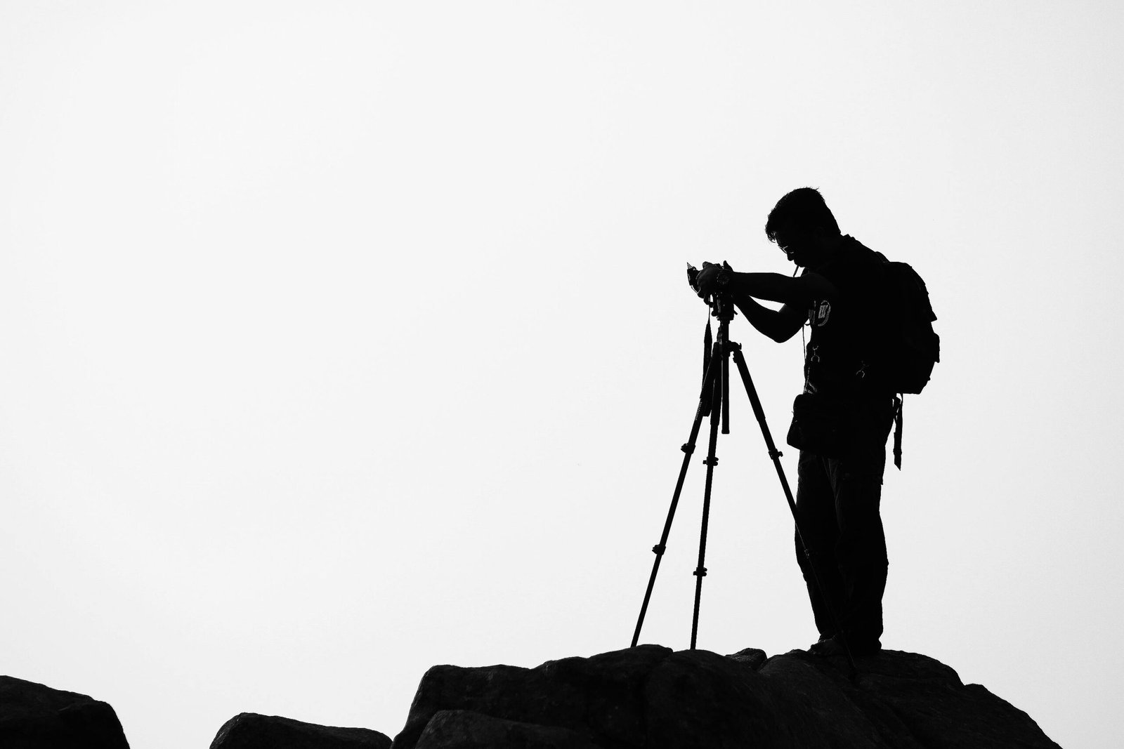 A photographer with a backpack uses a tripod camera on a cliff's edge during a cloudy day.